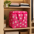 A bright pink travel storage bag with a white and yellow daisy floral pattern sits on a wooden shelf inside an open closet, surrounded by folded clothes and small plants.